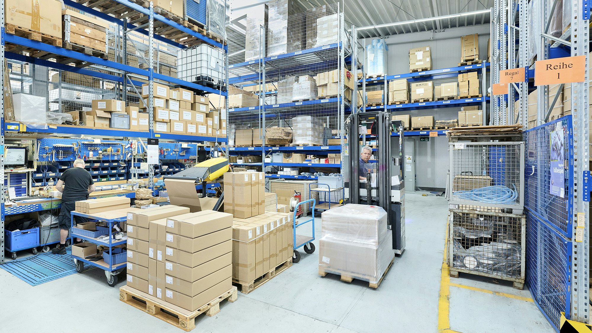 Palettenregale, in der Mitte eine Werkbank an der ein Mitarbeiter steht, während ein anderer Mitarbeiter auf einem Flurförderzeug eine Palette transportiert.