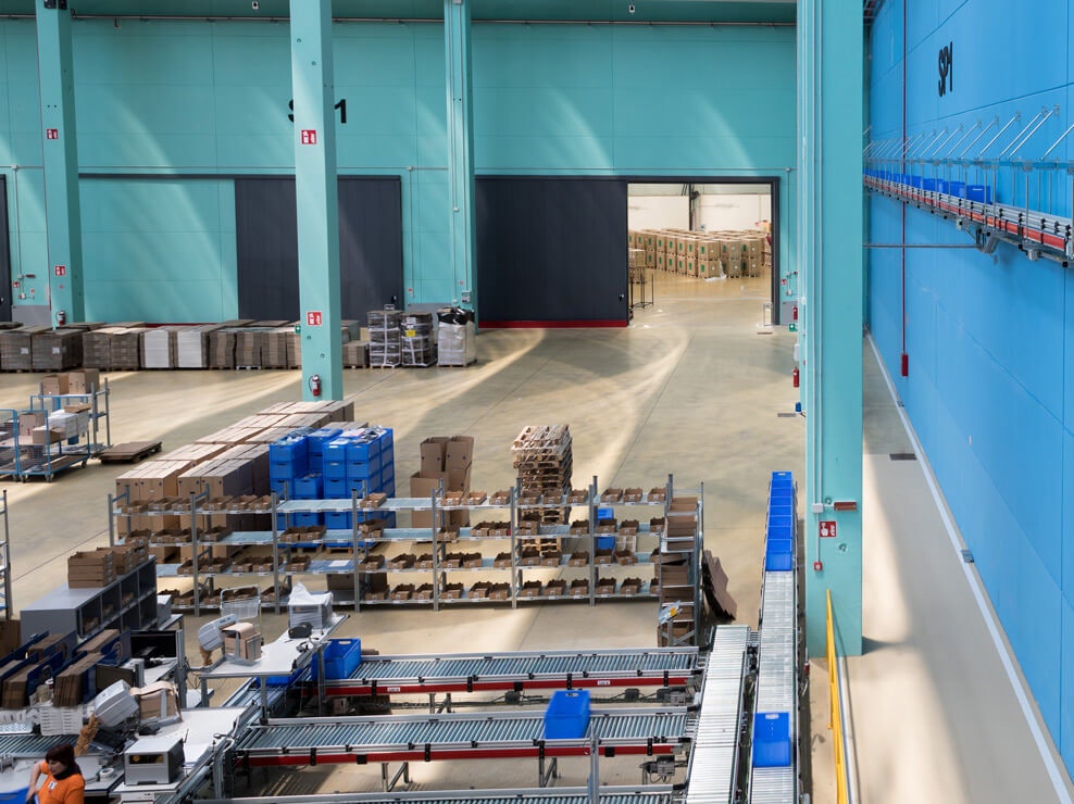 Eine Halle mit Rollenbahnen und Fachbodenregalen. An der Wand führt ein Förderband entlang.