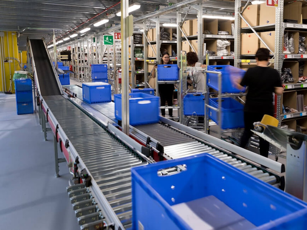 Vier Personen die, zwischen einer Rollenbahn und Fachbodenregalen, BITO Eurostapelbehälter auf Rollwägen transportieren.