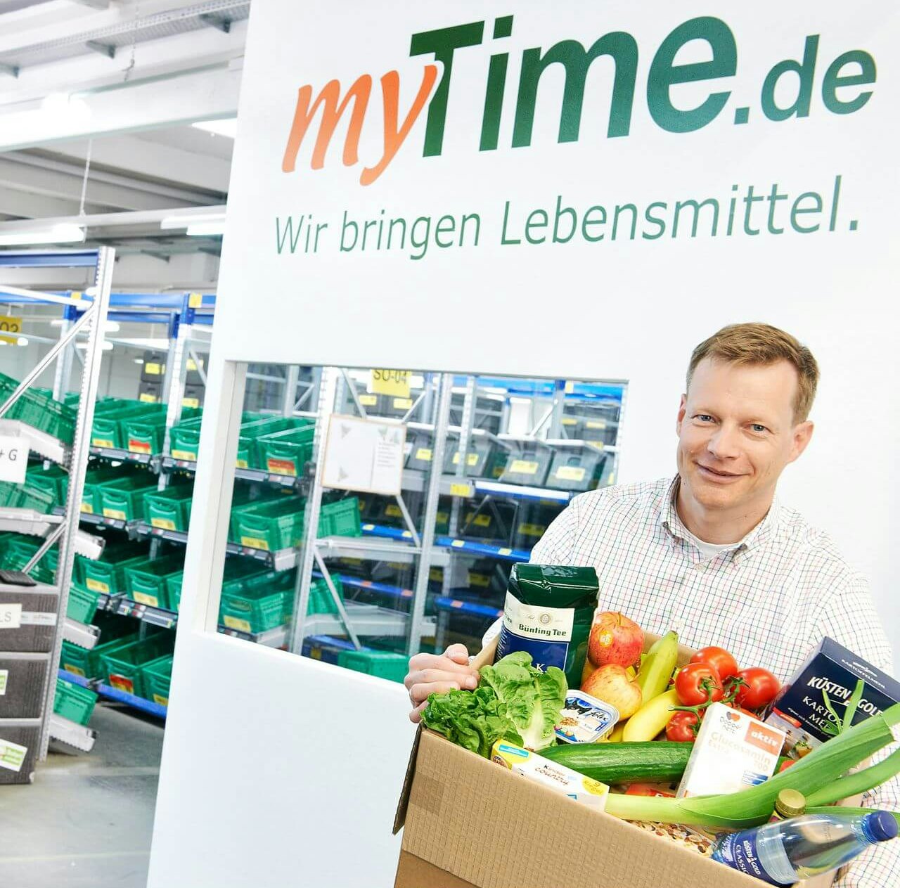 Ein Mann mit Lebensmitteln in einem Karton vor einem Werbebanner. Im Hintergrund BITO Fachbodenregale.