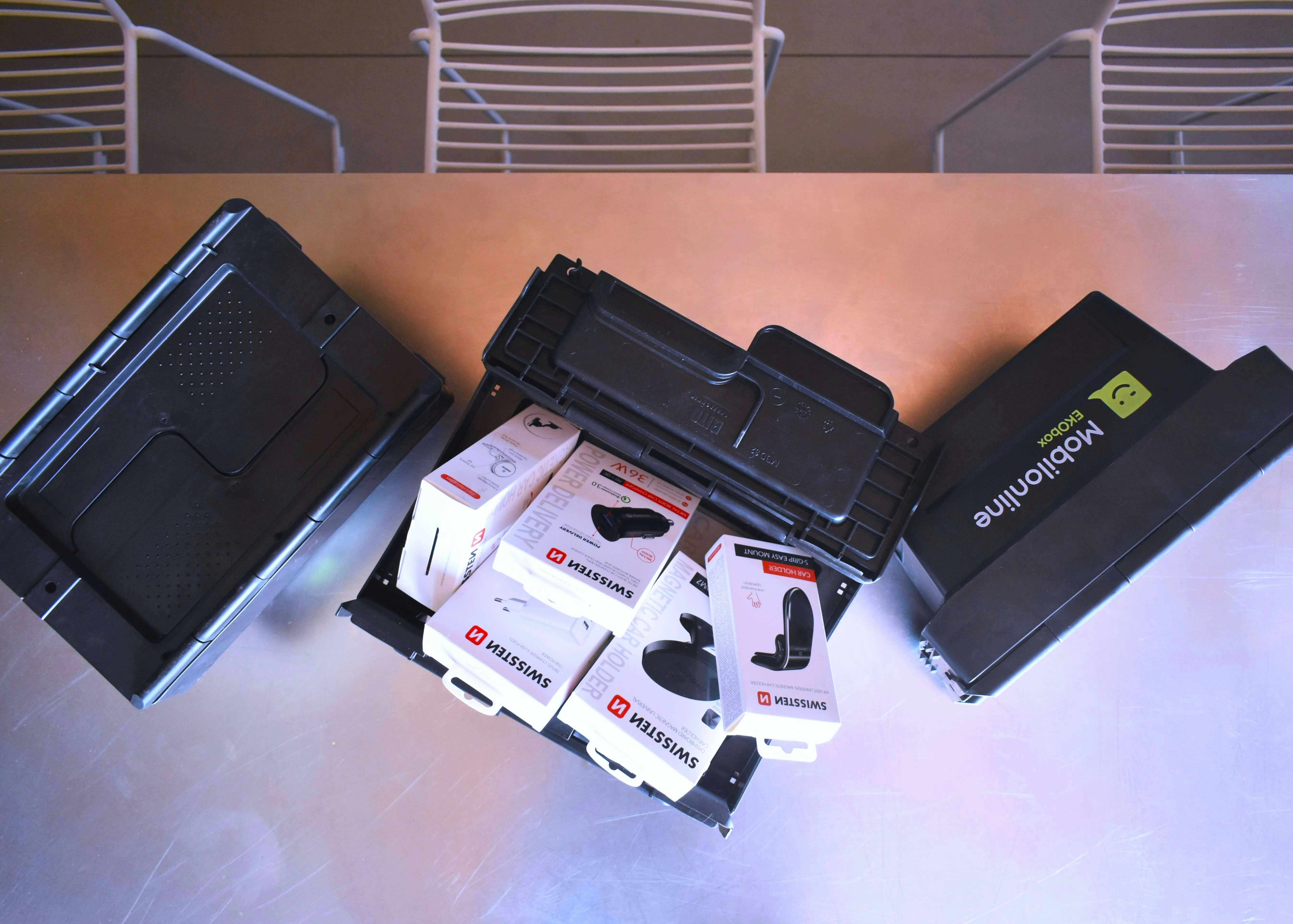 Drei schwarze BITO Behälter, einer mit der Aufschrift Mobileonline werden von oben gezeigt. Der Mittelste ist mit Produkten von Swissten befüllt.