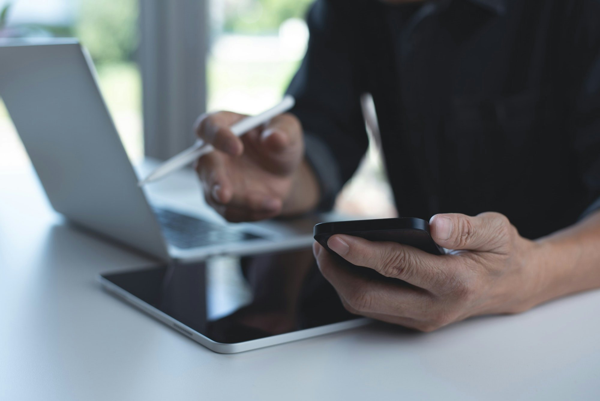 Nahaufnahme von den Händen eines Mannes. In der einen Hand hält er seinen Ipad Stift und in der Anderen sein Handy. Vor ihm auf dem Tisch liegt ein Ipad und steht ein Laptop.