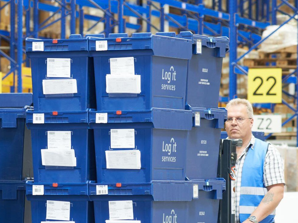 BITO Mehrwegbehälter MB Ein Mann transportiert gestapelte volumenreduzierte BITO Mehrwegbehälter mit der Aufschrift Log it services. Im Hintergrund befindet sich ein BITO Regal.