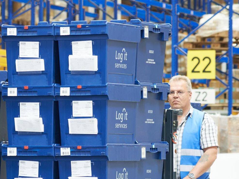 Pojemniki transportowe MB Ein Mann transportiert gestapelte volumenreduzierte BITO Mehrwegbehälter mit der Aufschrift Log it services. Im Hintergrund befindet sich ein BITO Regal.