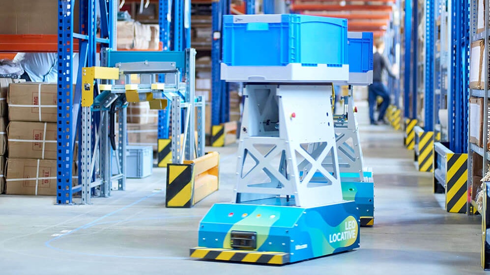 BITO LEO Ein LEO Locative fährt auf seiner Spur zwischen BITO Regalen in Richtung Übergabestation. Er transportiert einen BITO Behälter.