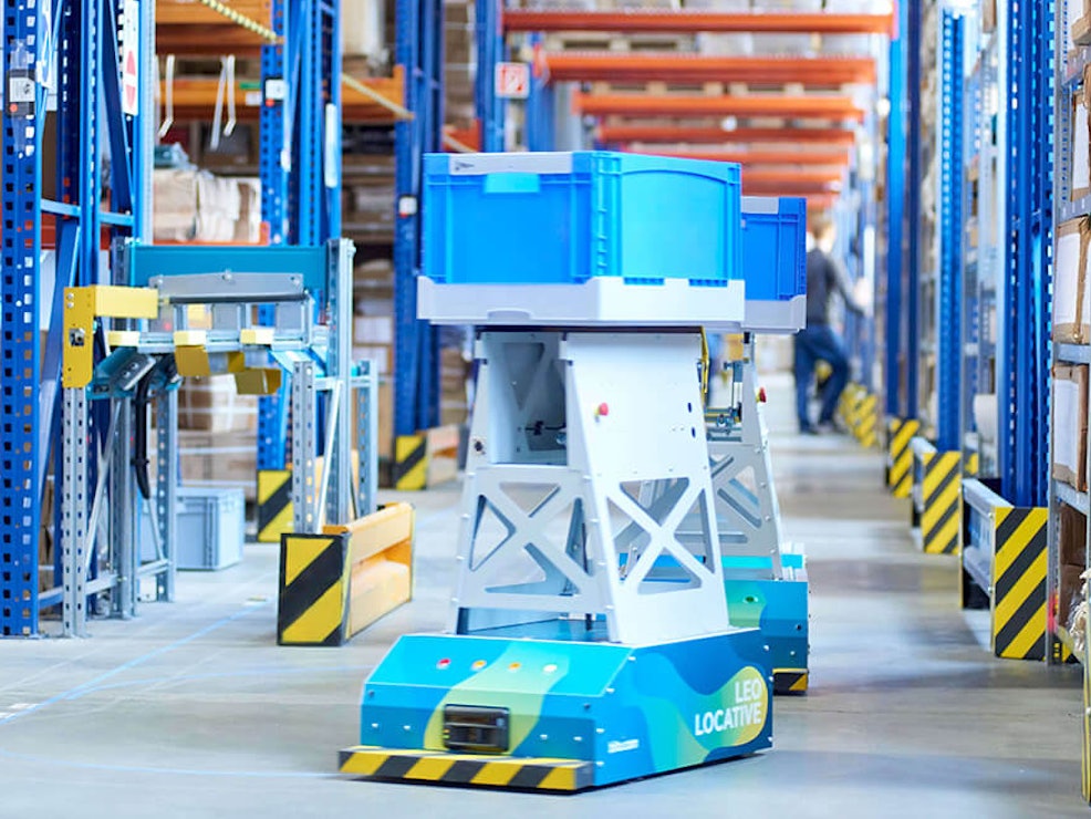 BITO LEO Ein LEO Locative fährt auf seiner Spur zwischen BITO Regalen in Richtung Übergabestation. Er transportiert einen BITO Behälter.