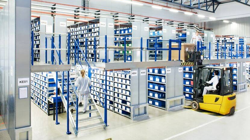 BITO Mehrgeschossanlage Eine BITO Lagerlösung auf zwei Etagen. Eine Frau geht gerade die Treppe nach oben, während rechts von ihr ein Paket mit dem Stapler in die zweite Etage gelagert wird.