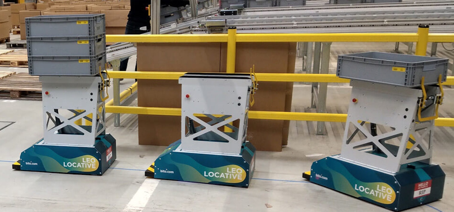Véhicules BITO LEO Drei LEO Locative fahren auf ihrer Bahn in einer Halle. Zwei davon sind mit Behälter beladen. Sie bewegen sich in Richtung der Übergabestation von BITO.