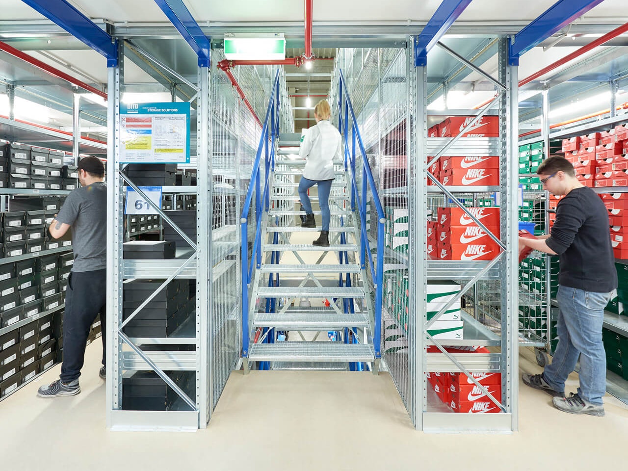 Wielopiętrowe instalacje regałowe i regały półkowe BITO Eine Mehrgeschossanlage wird als Schuhlager genutzt. In der Mitte läuft eine Frau über eine Treppe zur nächsten Etage. An den Regalen links und rechts daneben suchen zwei Mitarbeiter Schuhkartons aus dem Regal.