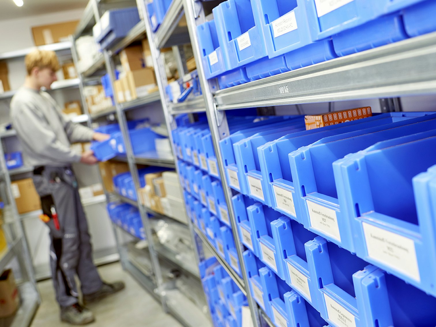 Regały z pojemnikami SK Kleine BITO Behälter für die Kleinteilelagerung in einem BITO Regal. Ein Mitarbeiter sucht sich Kleinteile aus einem Behälter.