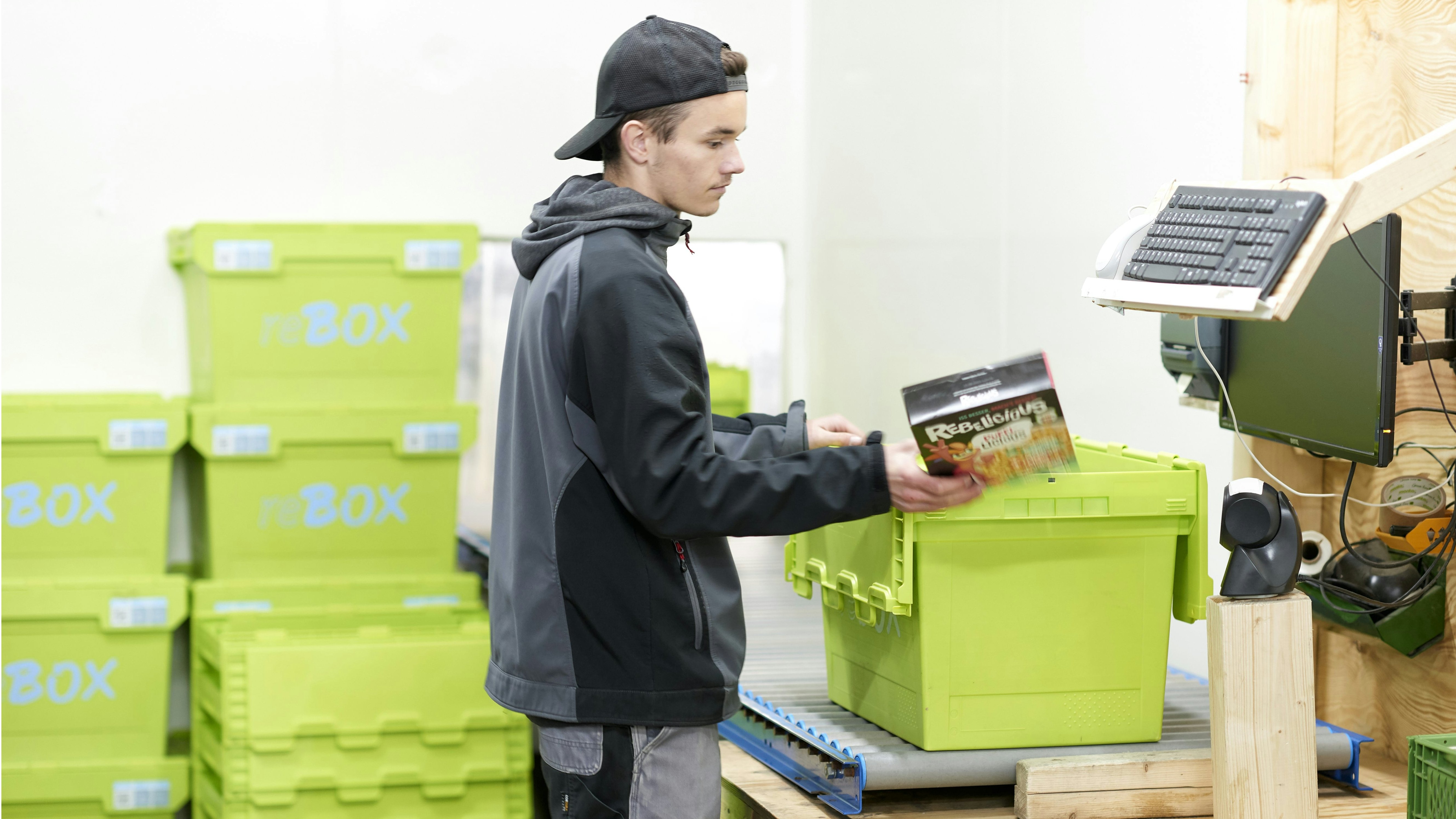 BITO Genanvendelig transportkasse MB Ein junger Mann räumt Lebensmittel aus einem BITO Behälter, der auf einem Förderband steht, aus. Im Hintergrund sind gestapelte BITO Behälter zu sehen.