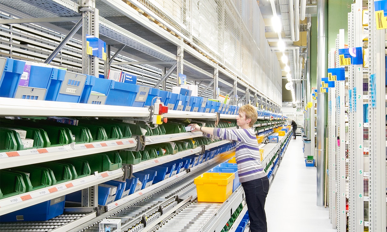 Eine Frau sortiert Produkte, die aus einem BITO Behälter über das Förderband laufen, in einen BITO Behälter aus einem Palettenregal ein.