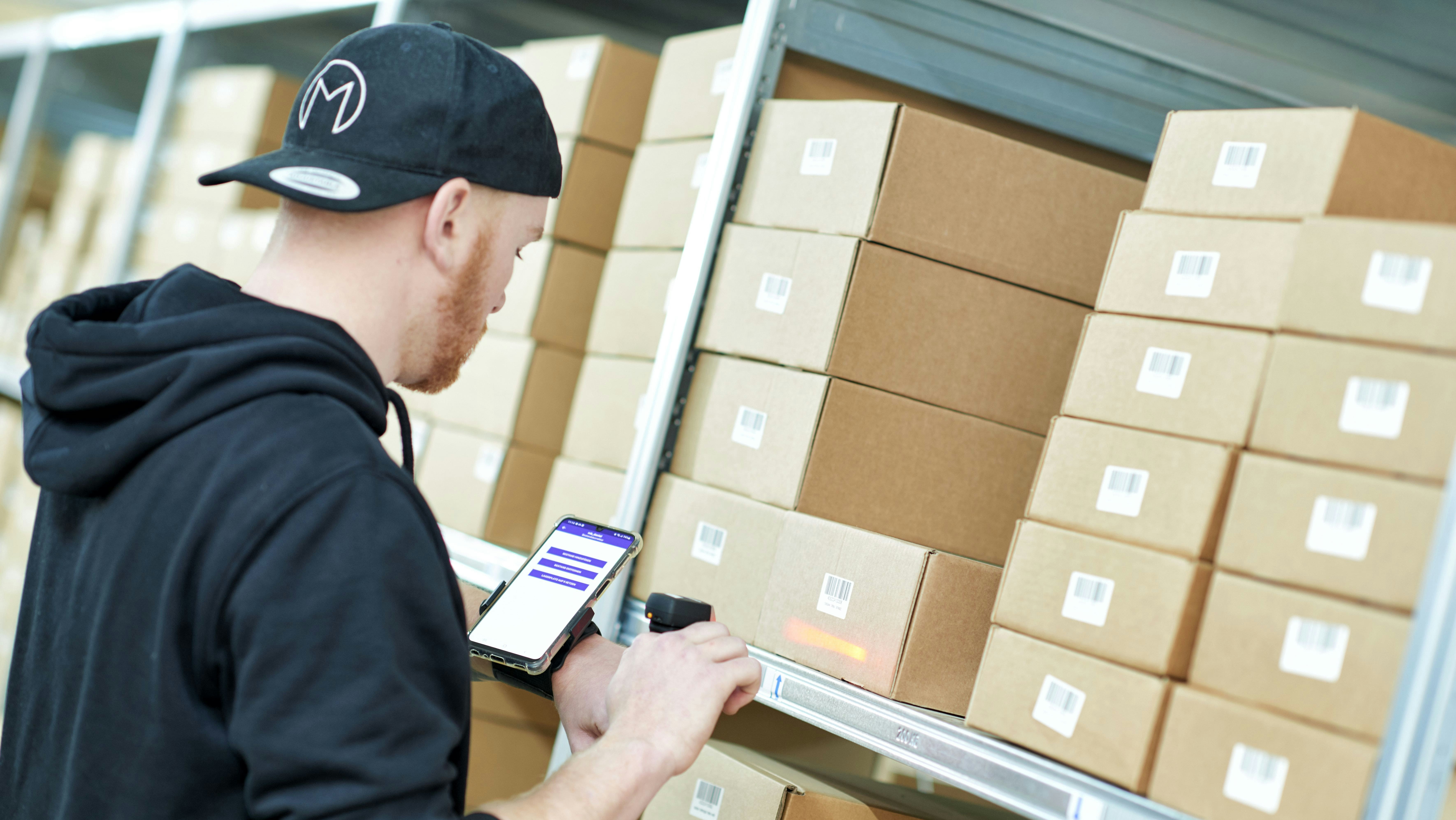 BITO WMS Ein Mann scannt Pakete, die in einem BITO Regal gestapelt sind. Die bereits gescannten Pakete werden ihm auf seinem Handy, das er um seinen Arm trägt, angezeigt.