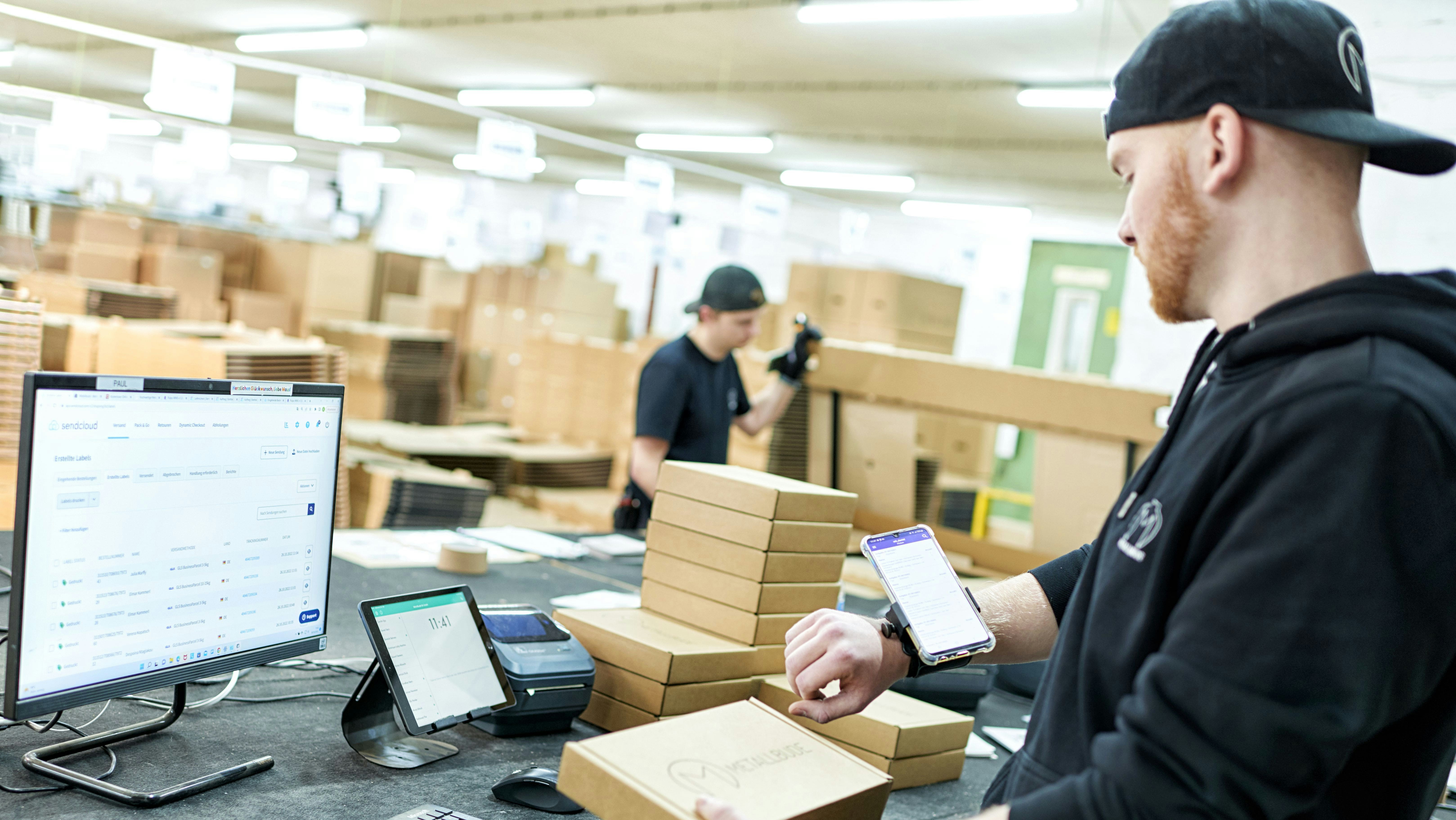 BITO WMS Ein Mann schaut sich auf seinem Handy gescannte Produkte an, während er in der anderen Hand ein Paket hält . Vor ihm steht ein Computer auf dem Tisch.