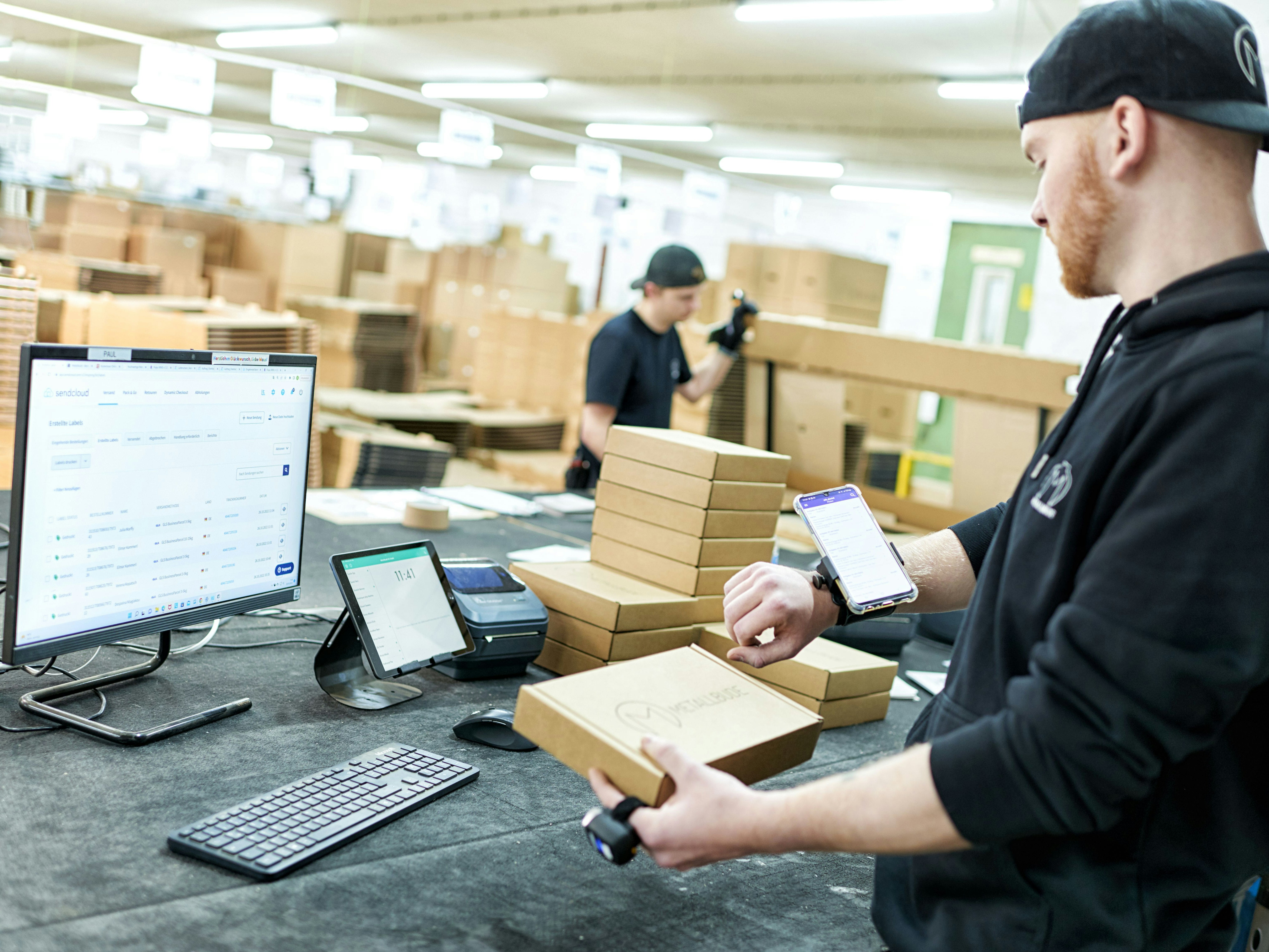 Rozwiązania do zarządzania magazynem BITO Ein Mann schaut sich auf seinem Handy gescannte Produkte an, während er in der anderen Hand ein Paket hält . Vor ihm steht ein Computer auf dem Tisch.