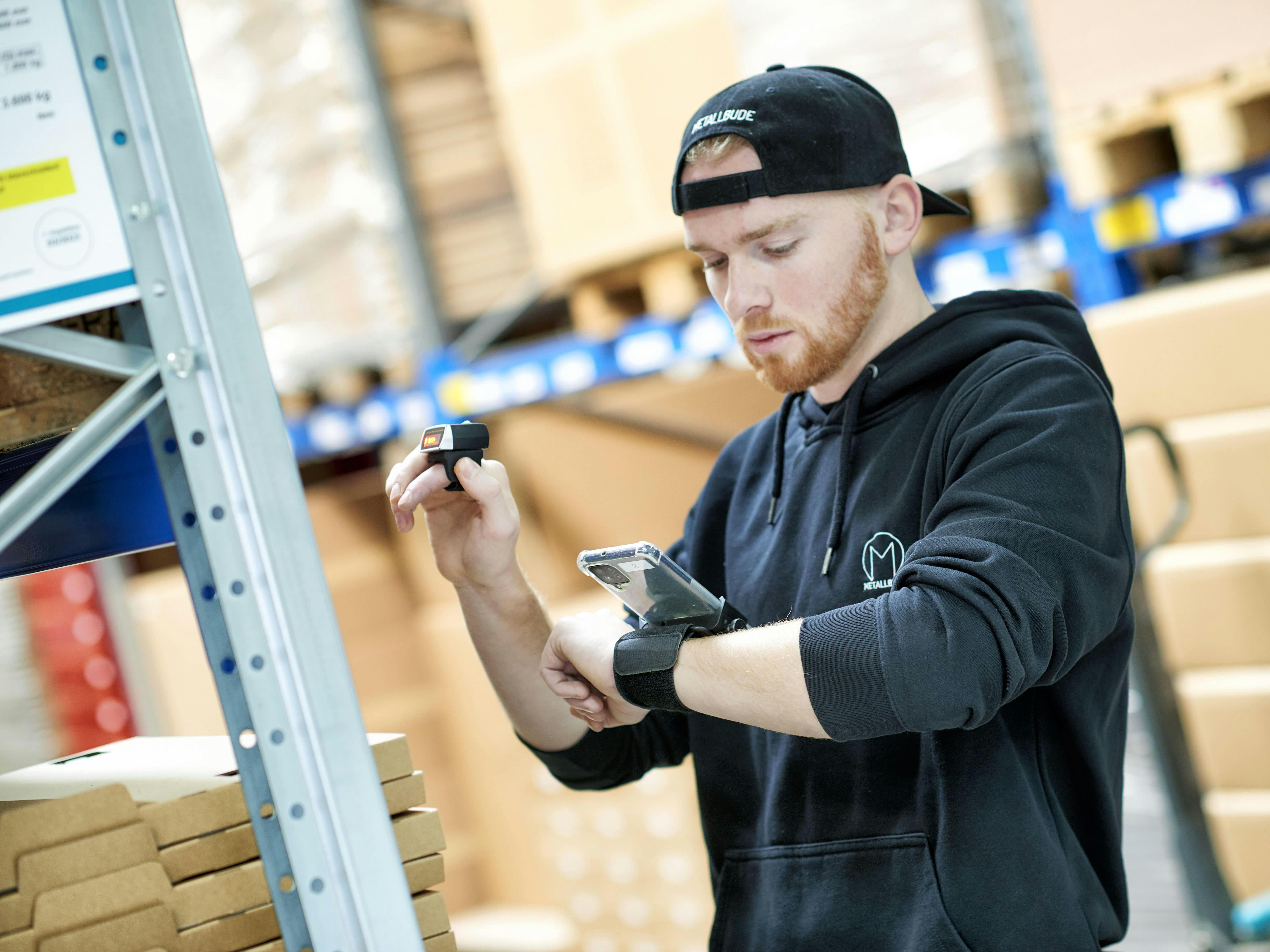 Ein Mann scannt Produkte aus einem BITO Regalsystem, welche auf seinem Handy, dass am linken Arm befestigt ist, angezeigt werden.