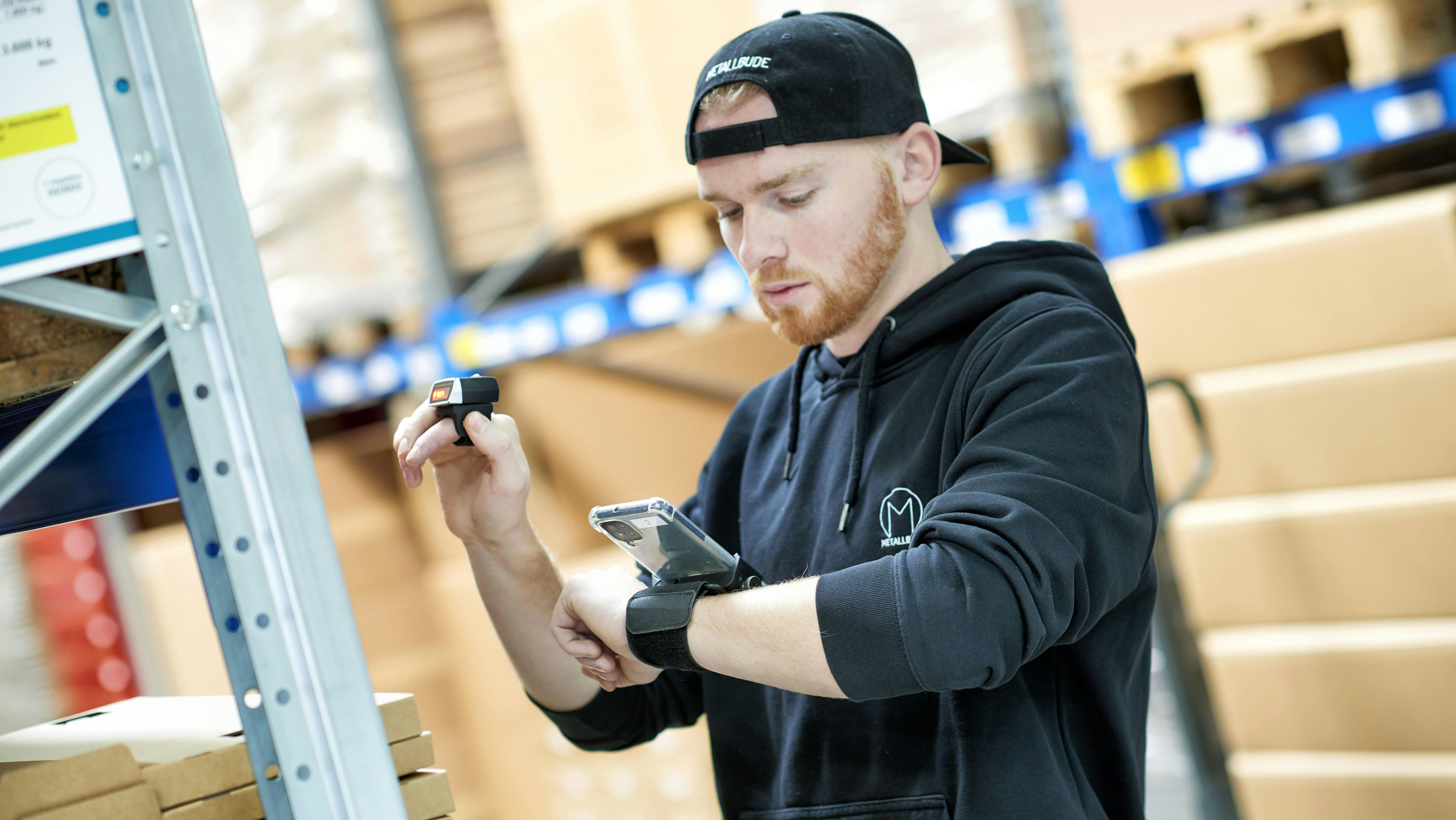 Ein Mann scannt Produkte aus einem BITO Regalsystem, welche auf seinem Handy, dass am linken Arm befestigt ist, angezeigt werden.