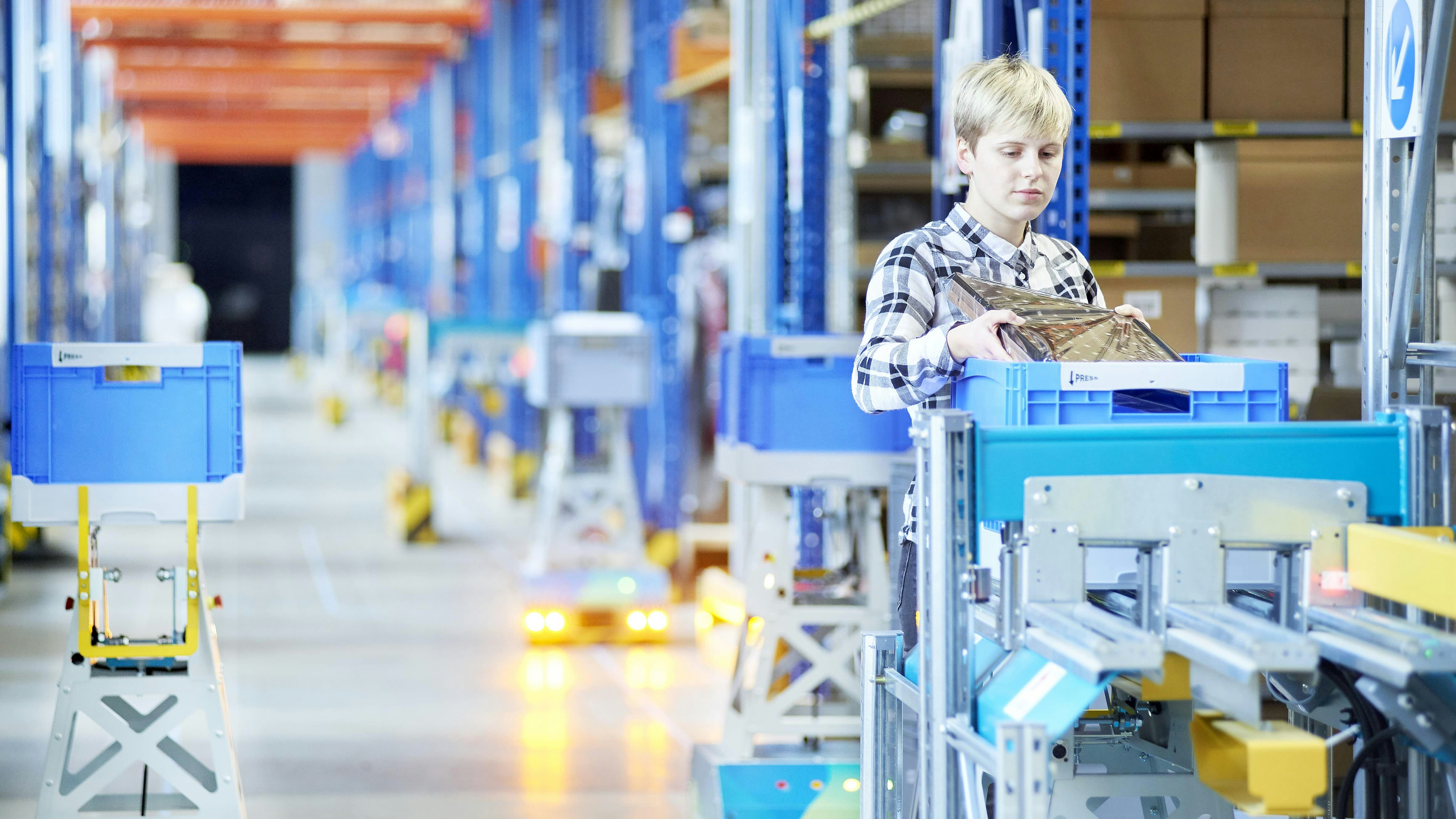BITO LEO Eine Frau legt ein Produkt in einen BITO Behälter, der auf der Übergabestation von LEO steht. Im Hintergrund fahren viele LEO Locative in einer Halle, die einen BITO Behälter transportieren.
