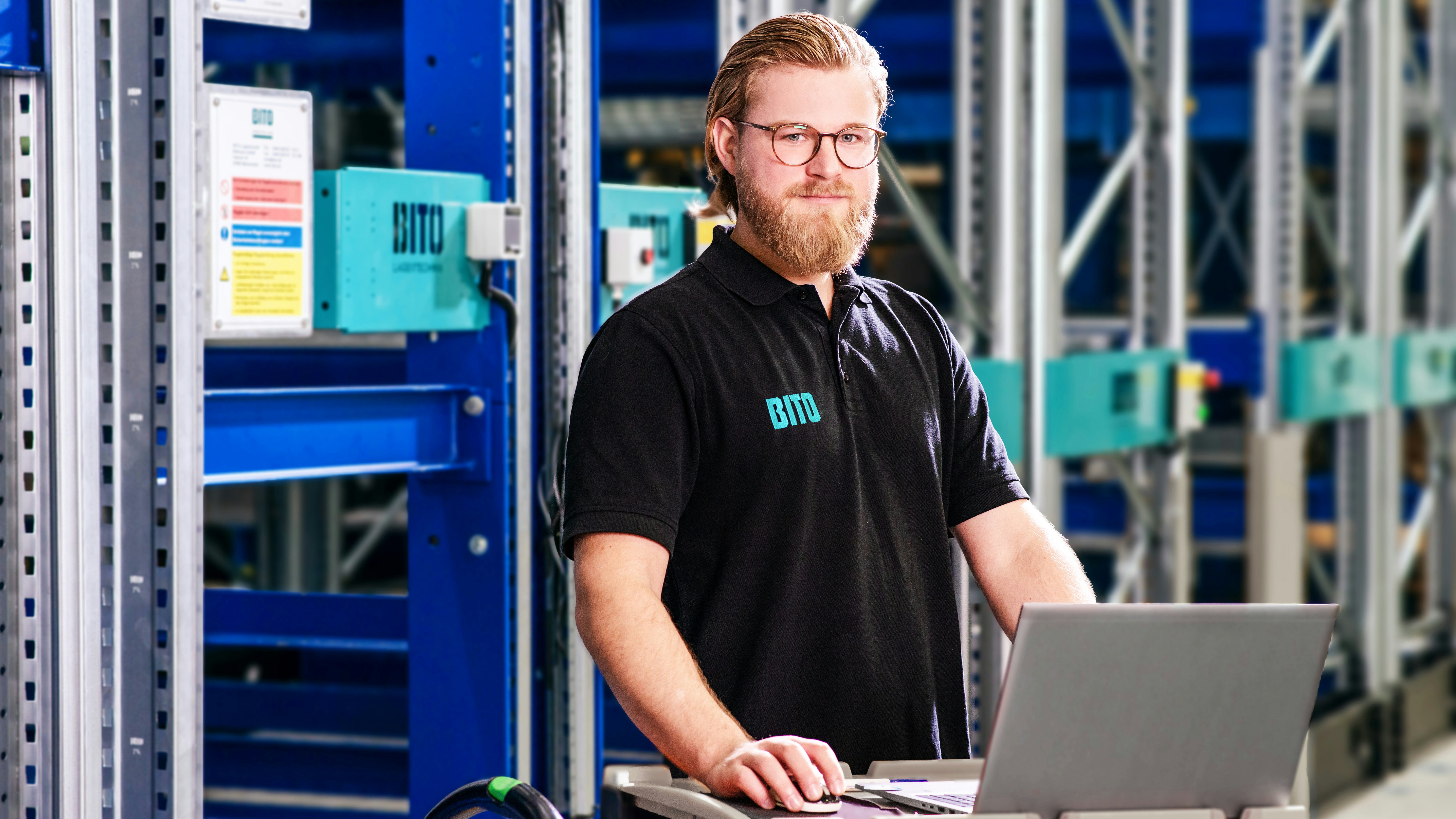 Ein lächelnder Mann mit einem Poloshirt von BITO steht vor einem BITO Lagerregal. Er hält eine Computermaus in der Hand und arbeitet an seinem Laptop, der auf gestapelten BITO Behältern vor ihm steht.
