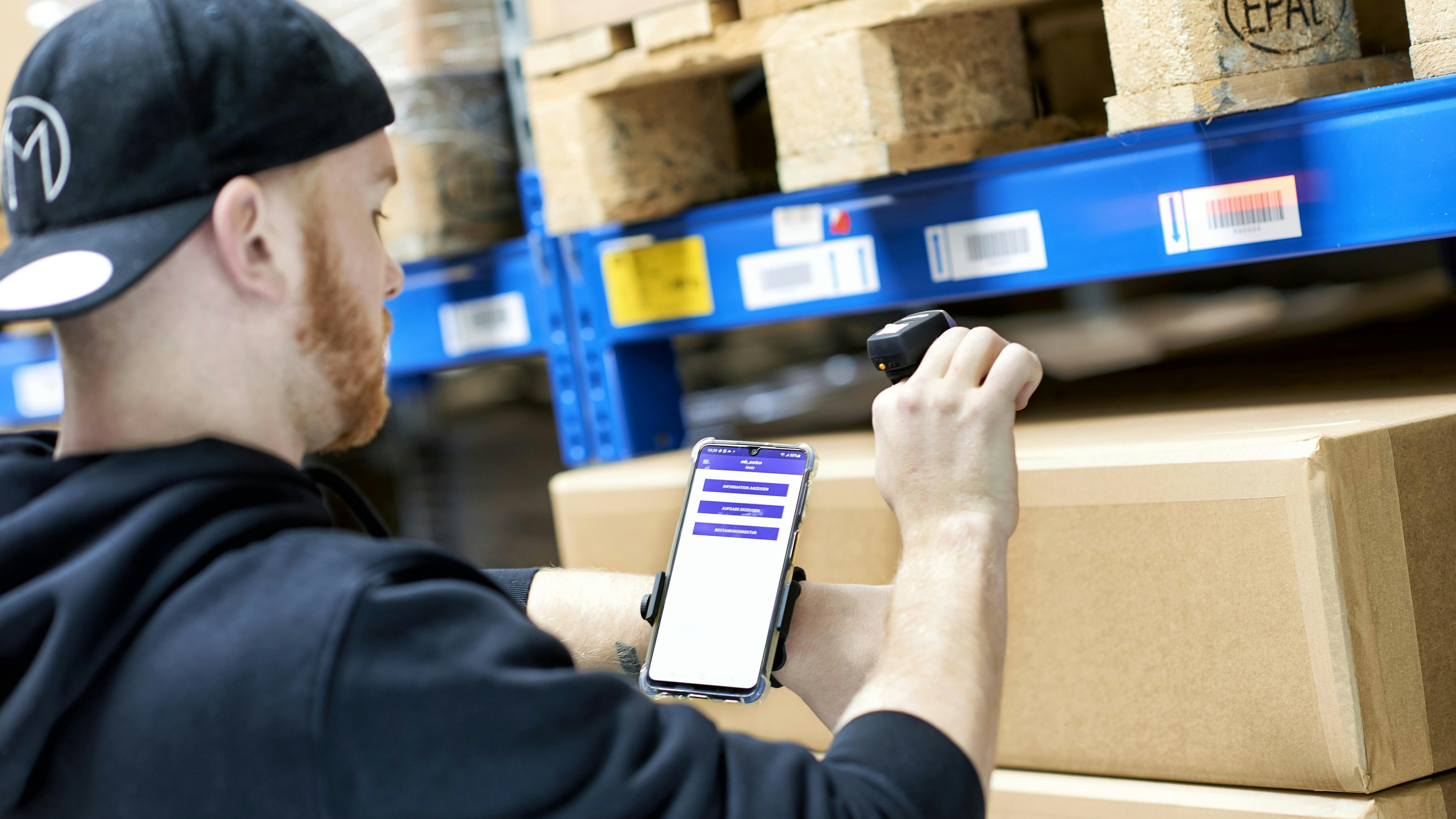 Logiciel de gestion d’entrepôt BITO Ein Mann wird von hinten fotografiert während er Produkte aus einem BITO Lagerregal abscannt und sich diese auf seinem Handy anguckt.