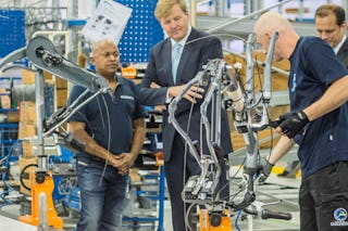 De foto toont Koning Alexander van Nederland die aan de montagelijn staat bij Gazelle fietsenbouwer en kijkt naar de montage van een fiets.