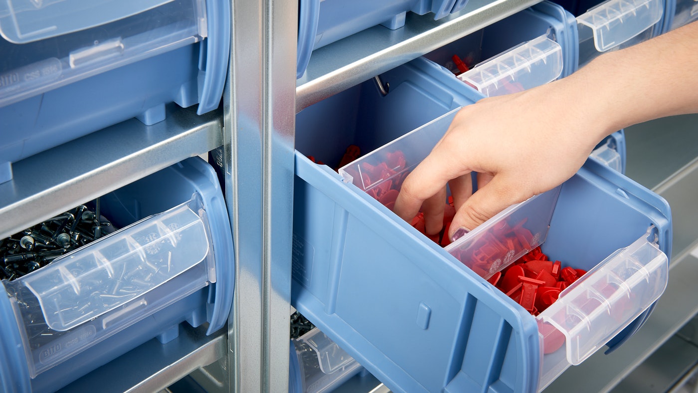 De foto toont een magazijnbak die uit een legbordstelling is getrokken en een hand van magazijnier neemt een klein c-onderdeel uit de bak