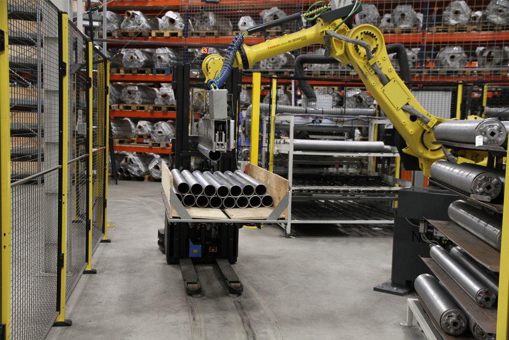 De foto toont een robotarm die lange buizen in lange houten kisten plaatst.