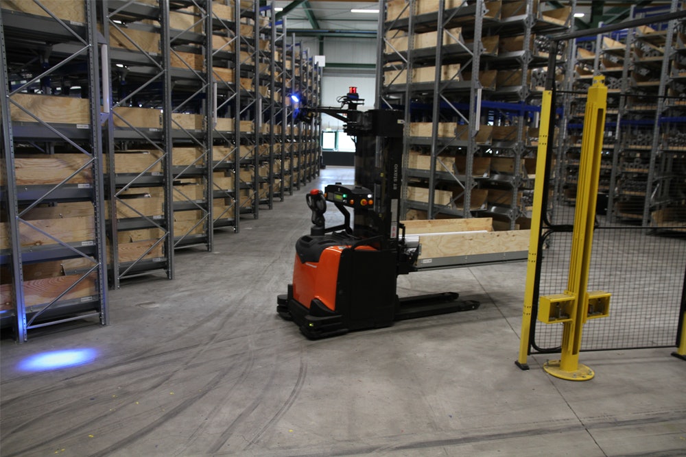 De foto toont een heftruck die rijdt tussen de inrijstellingen met een kist vol lange buizen