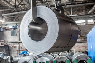 Ein Stahlcoil wird mit Hilfe eines Kranes abgeladen. Im Hintergrund stehen schon einige Coils im Lager