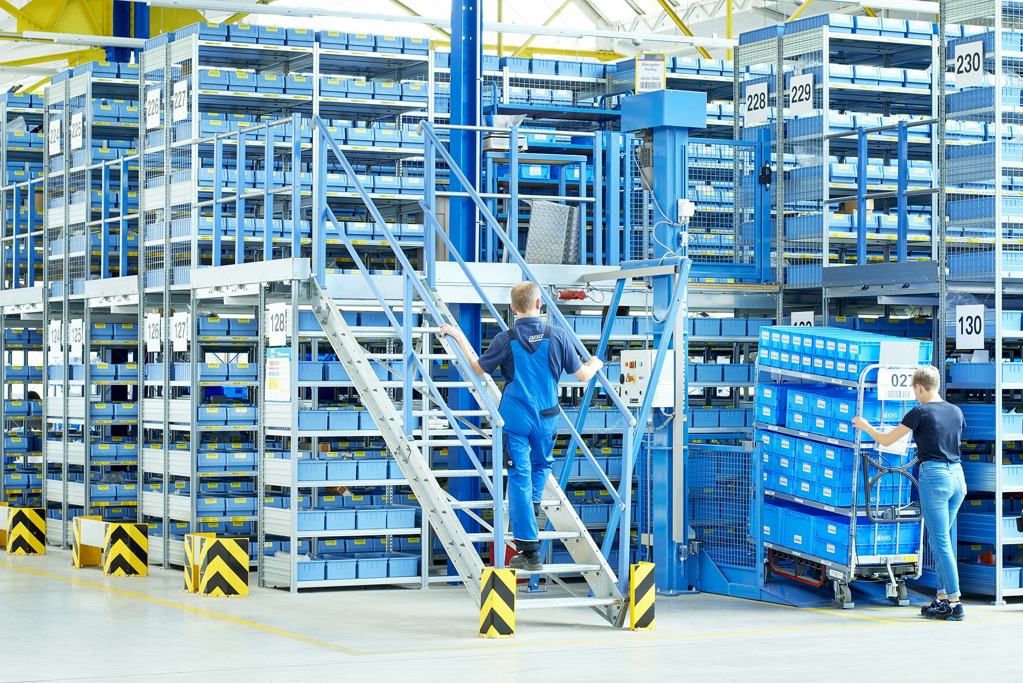 Ein Arbeiter geht auf einer Treppe in Richtung 2. Etage des Lagerregals, welches mit Behältern gefüllt ist.